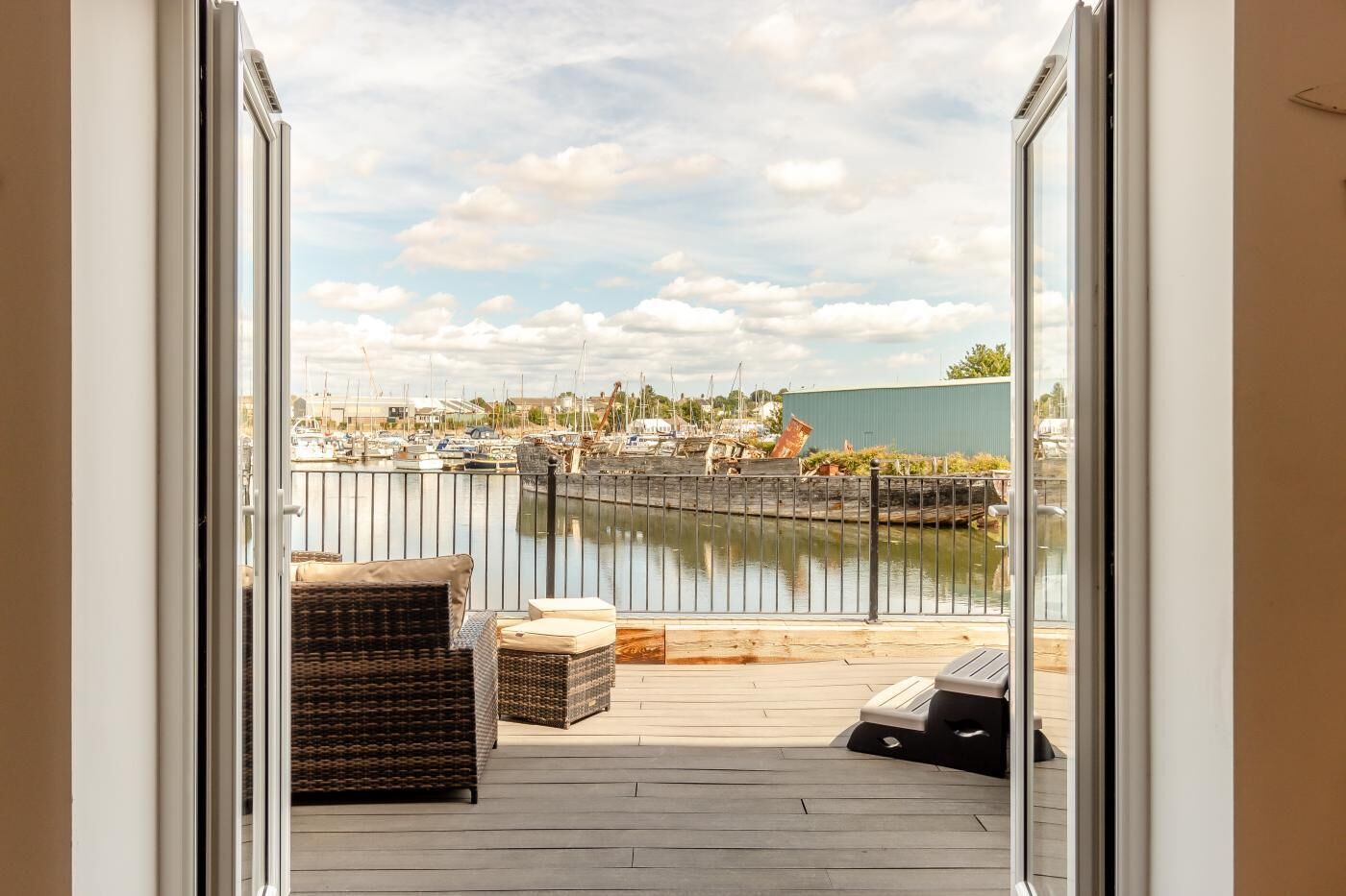 Een open deur die leidt naar een balkon met rieten meubels, uitzicht op een jachthaven met boten, en een groen gebouw op de achtergrond.