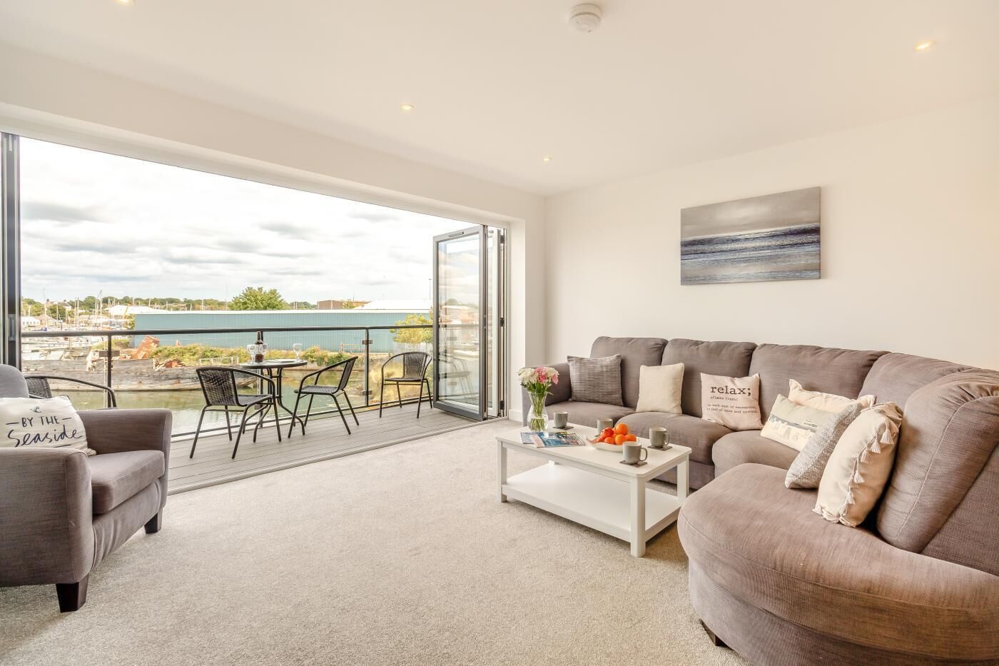 A modern living room with a large window, a grey sofa, a coffee table, and a balcony with outdoor seating.