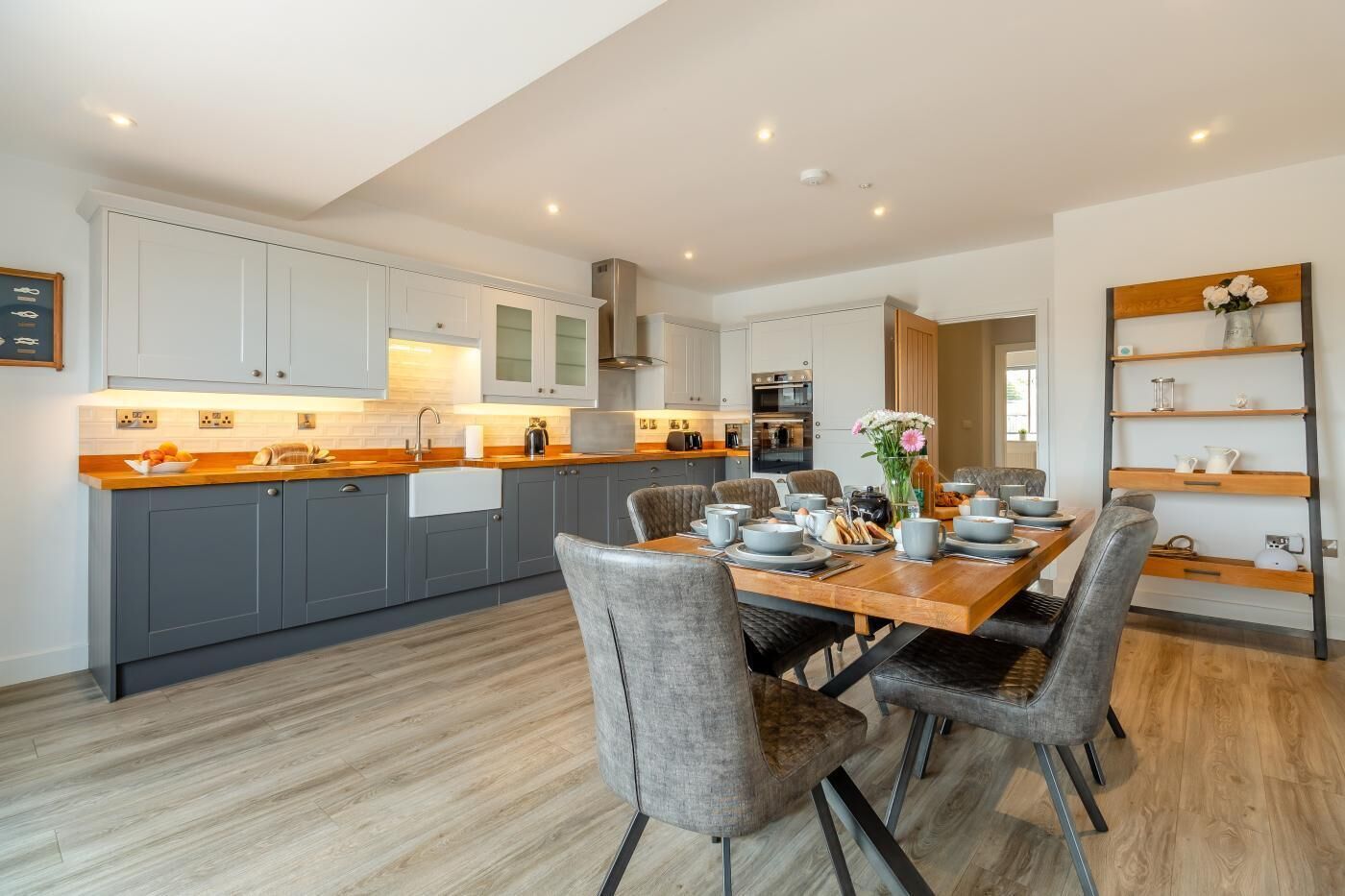 A modern kitchen with grey cabinets, a wooden countertop, and a dining area with grey upholstered chairs.