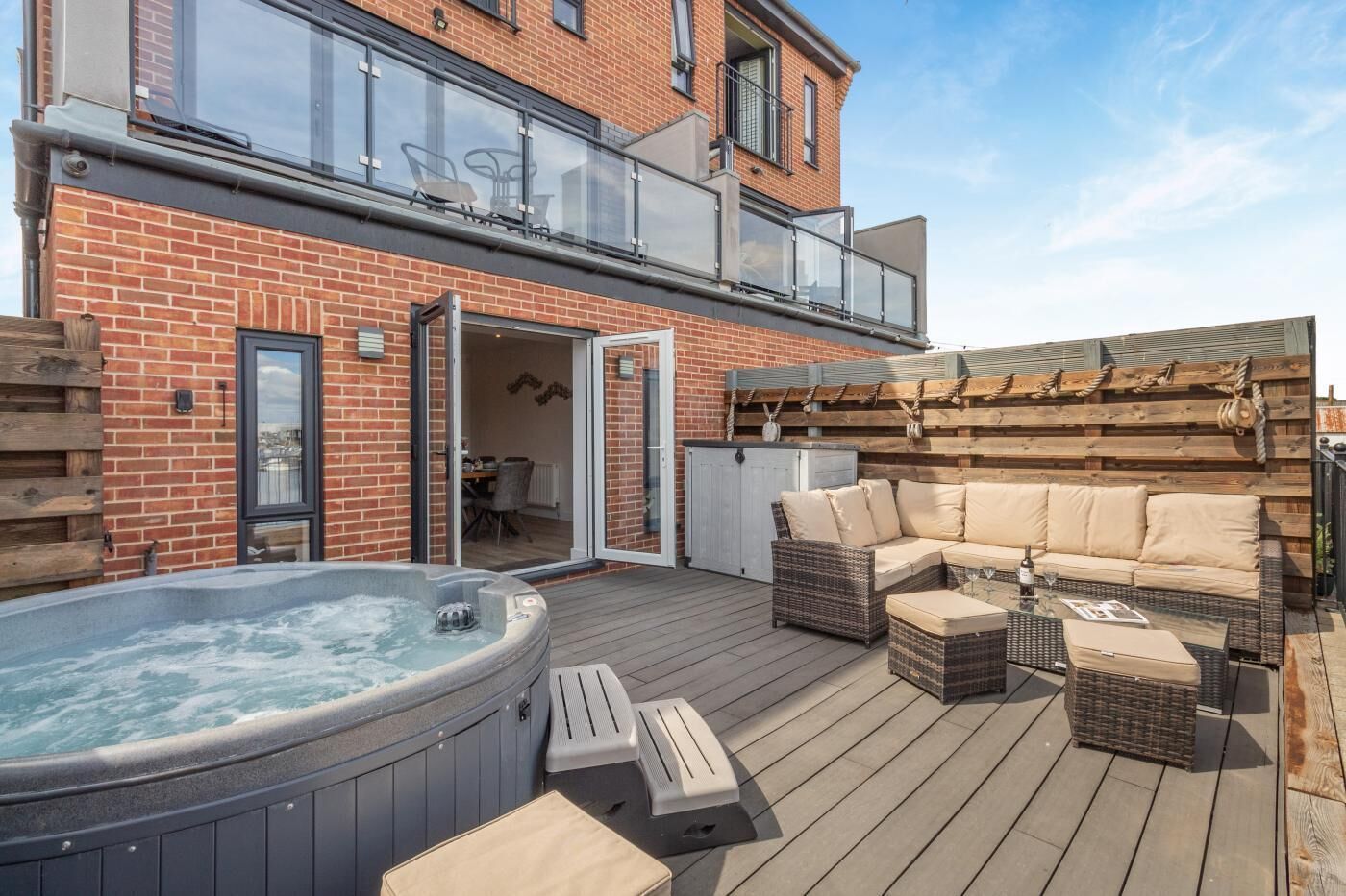 Eine Holzterrasse mit Whirlpool, Gartenmöbeln und einem Ziegelhaus mit Balkon.