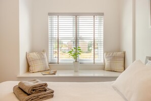 Ein Schlafzimmer mit großem Fenster, einem Bett mit Kissen, einem Nachttisch mit Pflanze und einem Stapel Handtücher.