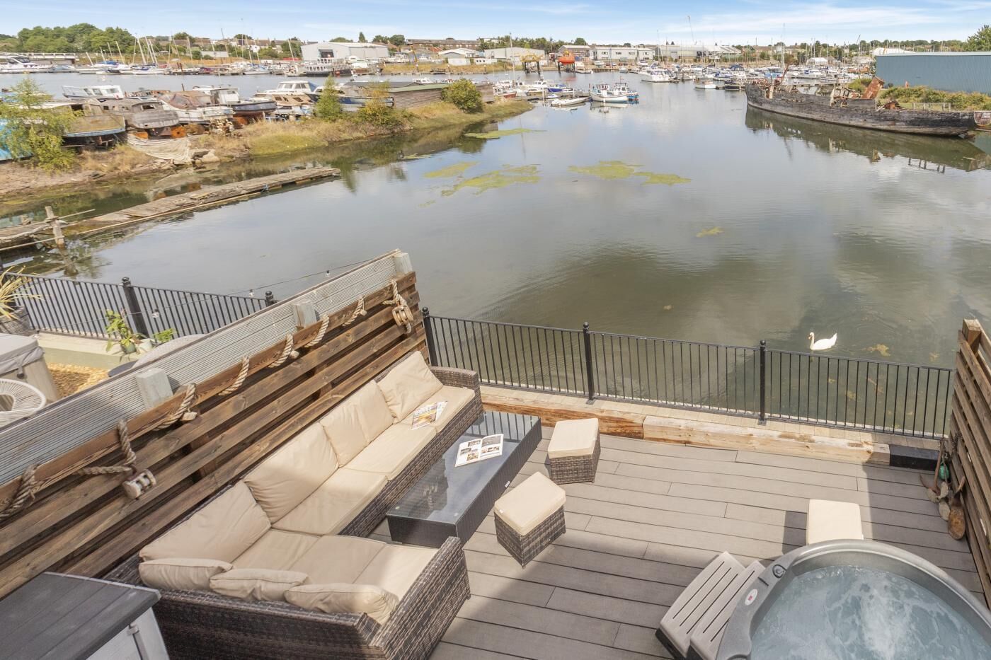 A deck with a sofa, a table, and a hot tub overlooking a calm water body with boats and buildings in the background.