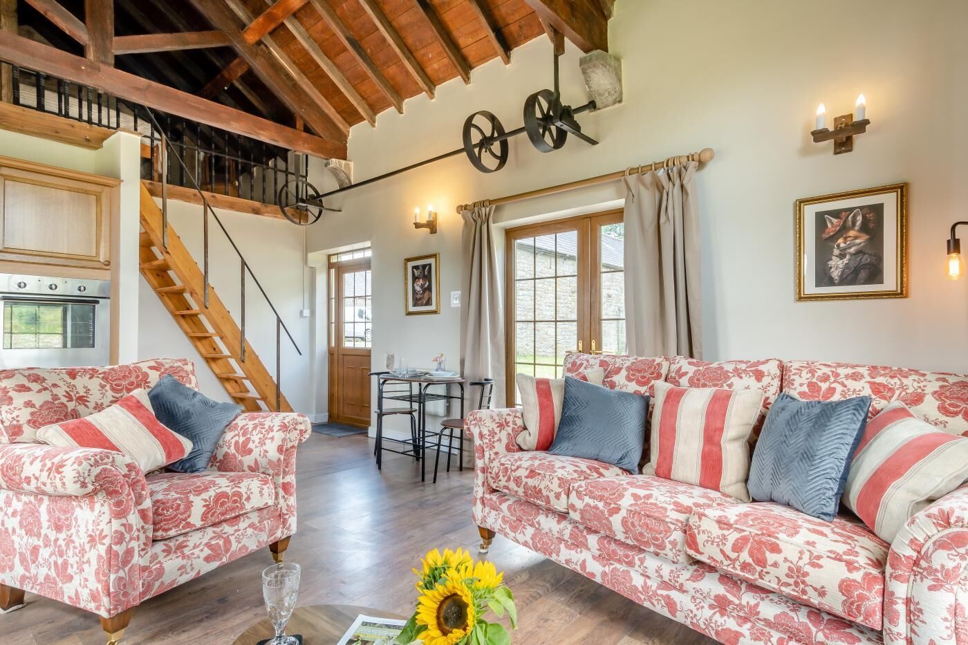 A cozy living room with floral-patterned sofas, a wooden staircase, and a dining area with a table and chairs.