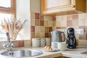Eine Küche mit Spüle aus Edelstahl, einer Kaffeemaschine, einem Croissant auf einem Teller und einer Vase mit Trockenblumen.