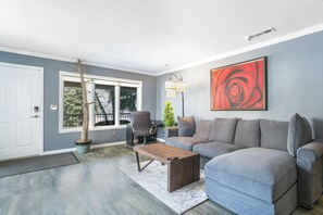Ein modernes Wohnzimmer mit einem grauen Sofa, einem Holztisch, einer Topfpflanze und einem großen gerahmten Kunstwerk.