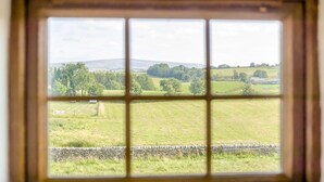Una ventana con vista a un paisaje rural, que incluye campos, árboles y un muro de piedra.