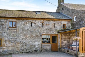 A stone building with a wooden door and windows, a covered porch with seating, and a small attached structure. - 2 Bed in Gilsland (oc-o45731) (Waterhead)