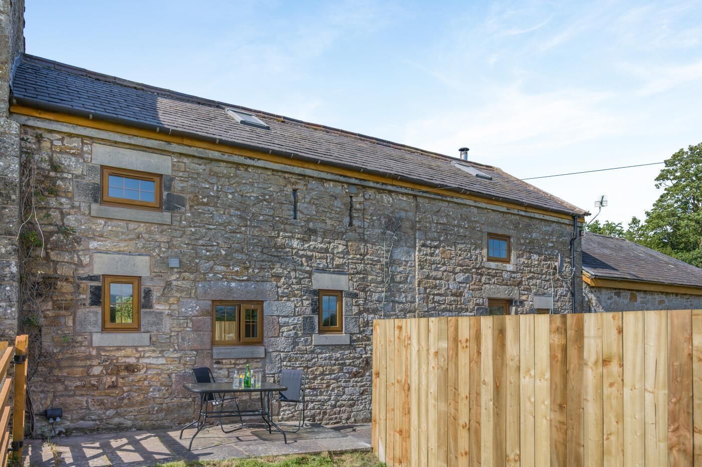A stone building with a wooden fence, a table with chairs, and a clear sky.