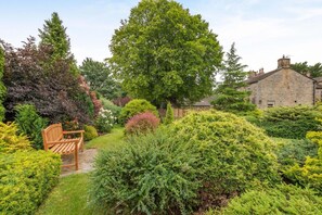 Ein Garten mit einer hölzernen Bank, verschiedenen Sträuchern und einem Steinhaus im Hintergrund.