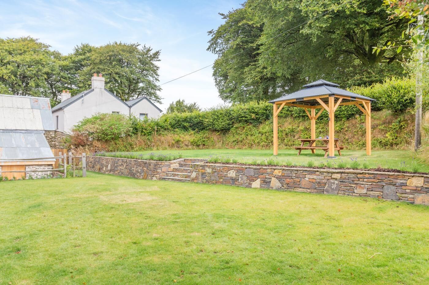 Un jardín con un muro de piedra, un pabellón de madera y una mesa de picnic.
