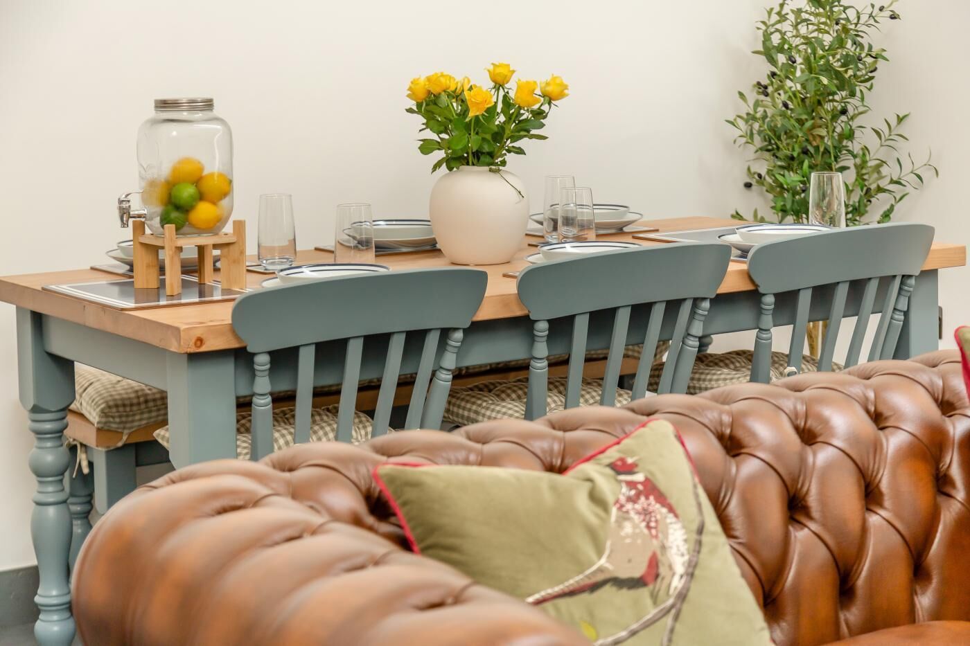 A dining table with chairs, a vase with yellow flowers, a glass jar with yellow balls, and a plant in the background.
