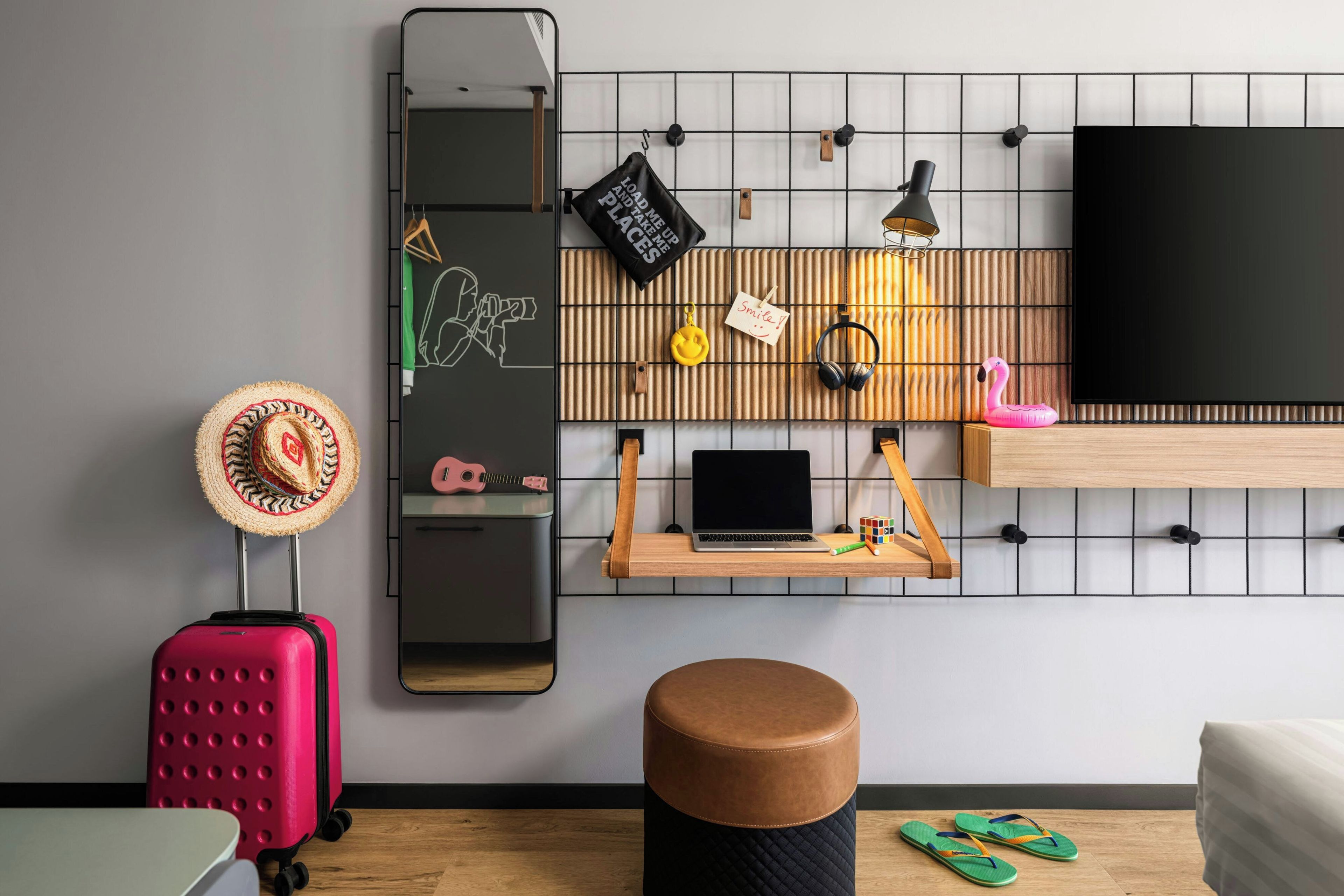 Ein modernes Zimmer mit einem großen Spiegel, einem Laptop auf einem Holzregal, einem pinken Koffer, einem Holzhocker und einem Paar grüner Badeschlappen.