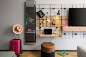 Una habitación moderna con un espejo grande, una laptop sobre una estantería de madera, una maleta rosa, un taburete de madera y un par de chanclas verdes.