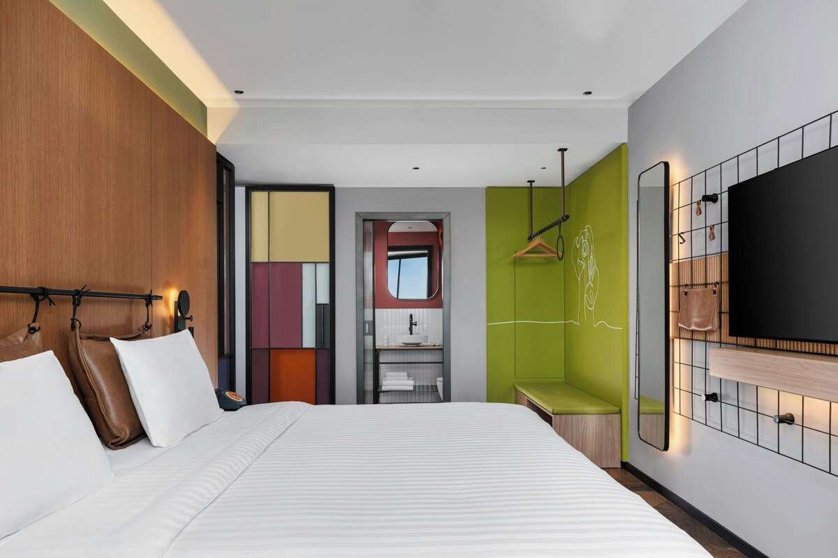 Habitación de hotel moderna con una cama grande, un televisor montado en la pared y un baño visible a través de una puerta abierta.