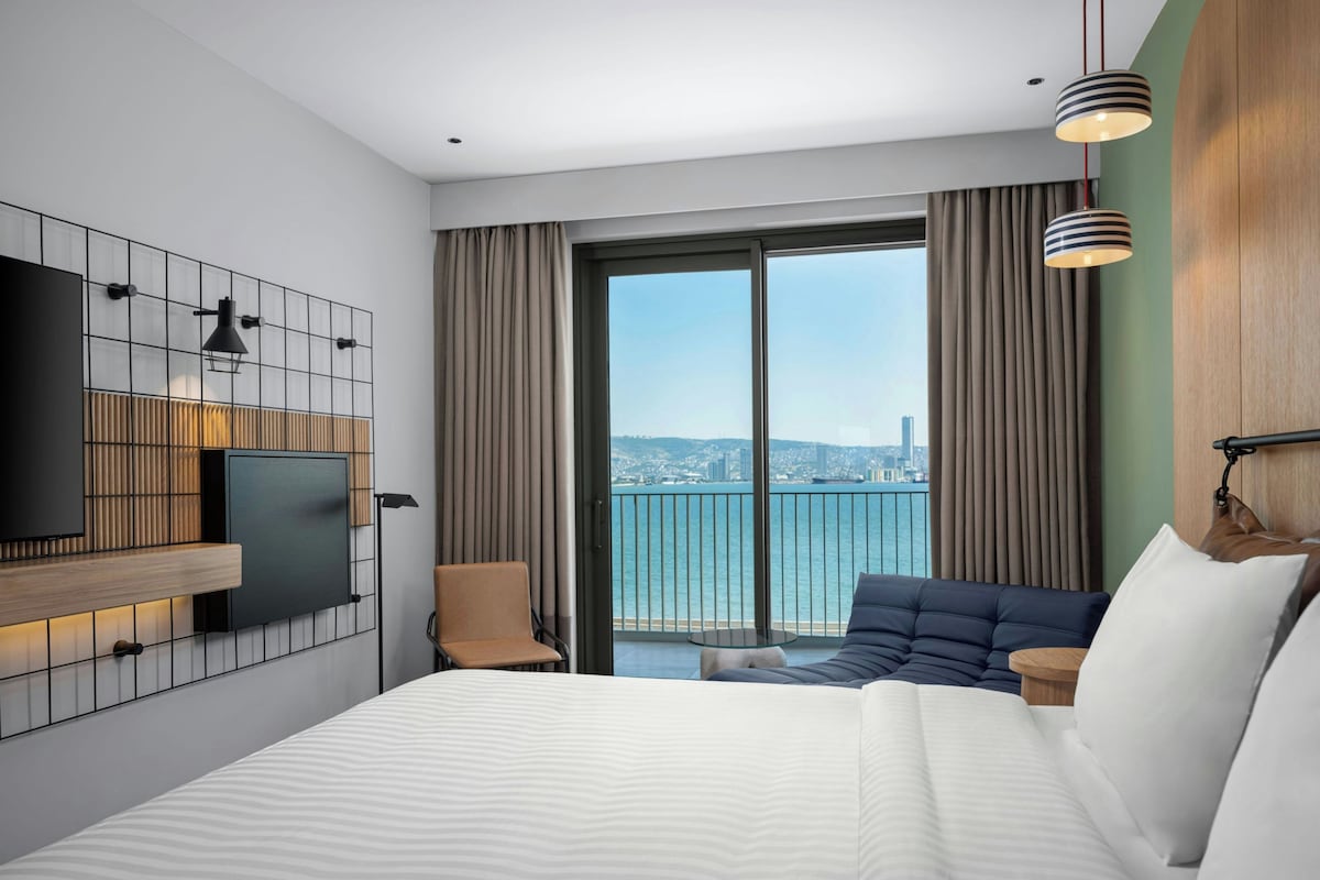 A modern hotel room with a large bed, a TV mounted on a wall, a balcony with a city view, and a cityscape visible through the window.