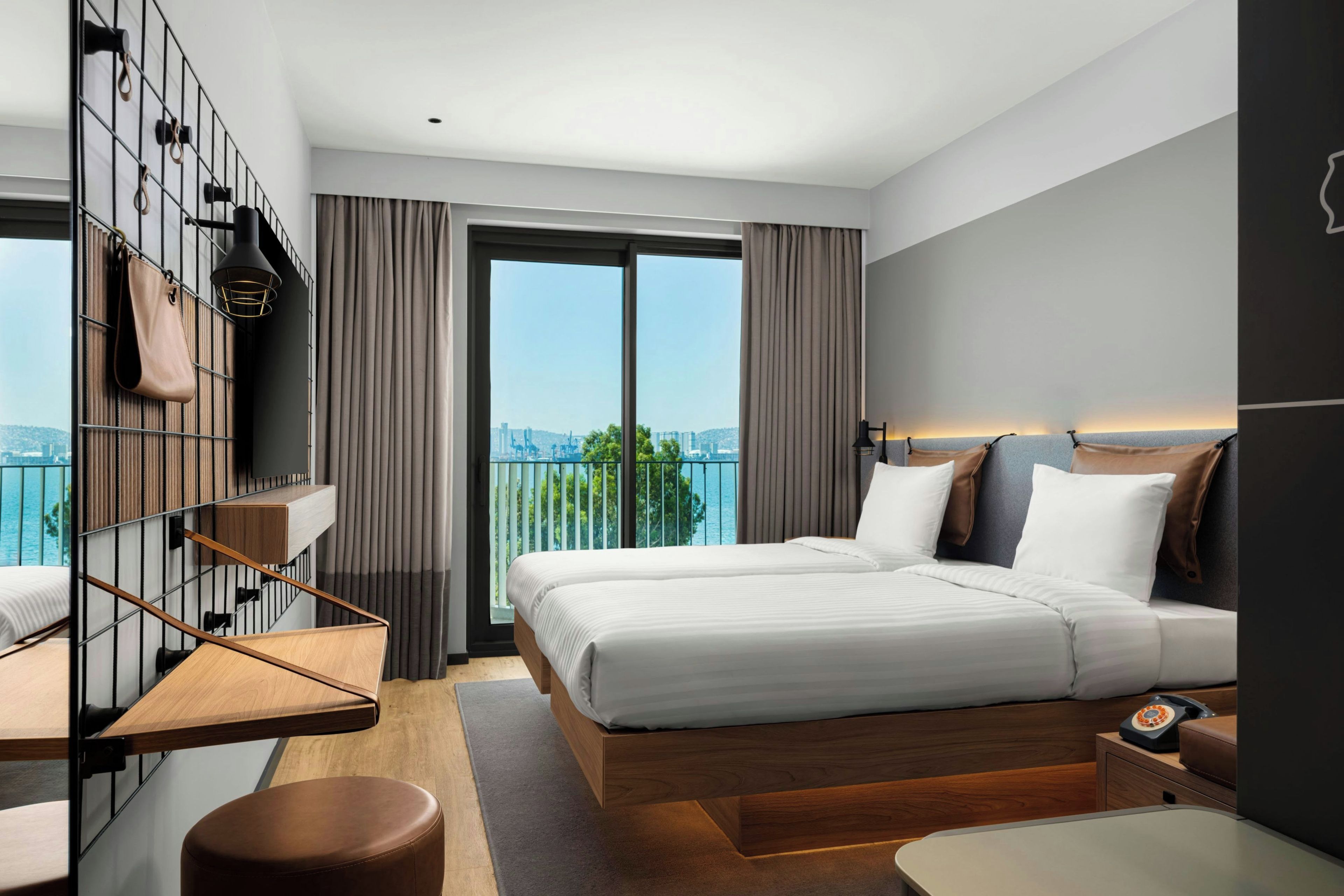 A modern hotel room with a large bed, a desk, a chair, a small table, a telephone, and a view of the city through the window.