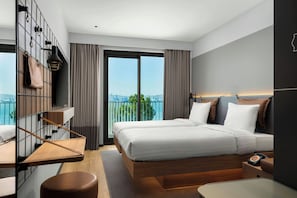A modern hotel room with a large bed, a desk, a chair, a small table, a telephone, and a view of the city through the window. - Moxy Izmir (İzmir)