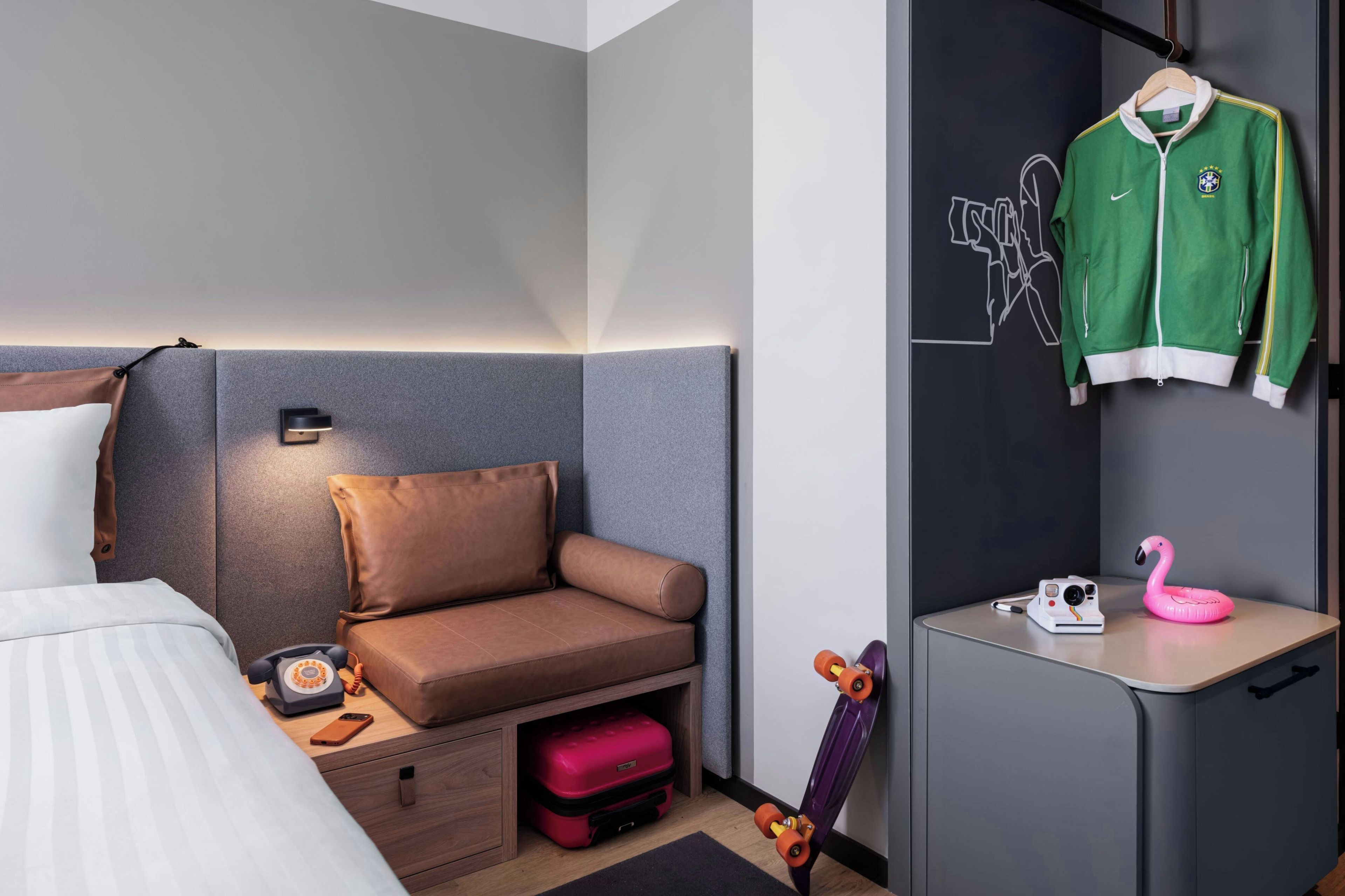 A modern hotel room with a bed, a sofa, a desk, a hanging jacket, a pink flamingo-shaped camera, and a skateboard.