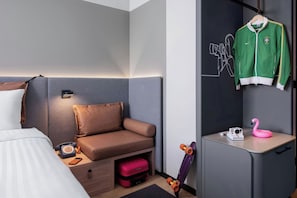 A modern hotel room with a bed, a sofa, a desk, a hanging jacket, a pink flamingo-shaped camera, and a skateboard. - Moxy Izmir (İzmir)