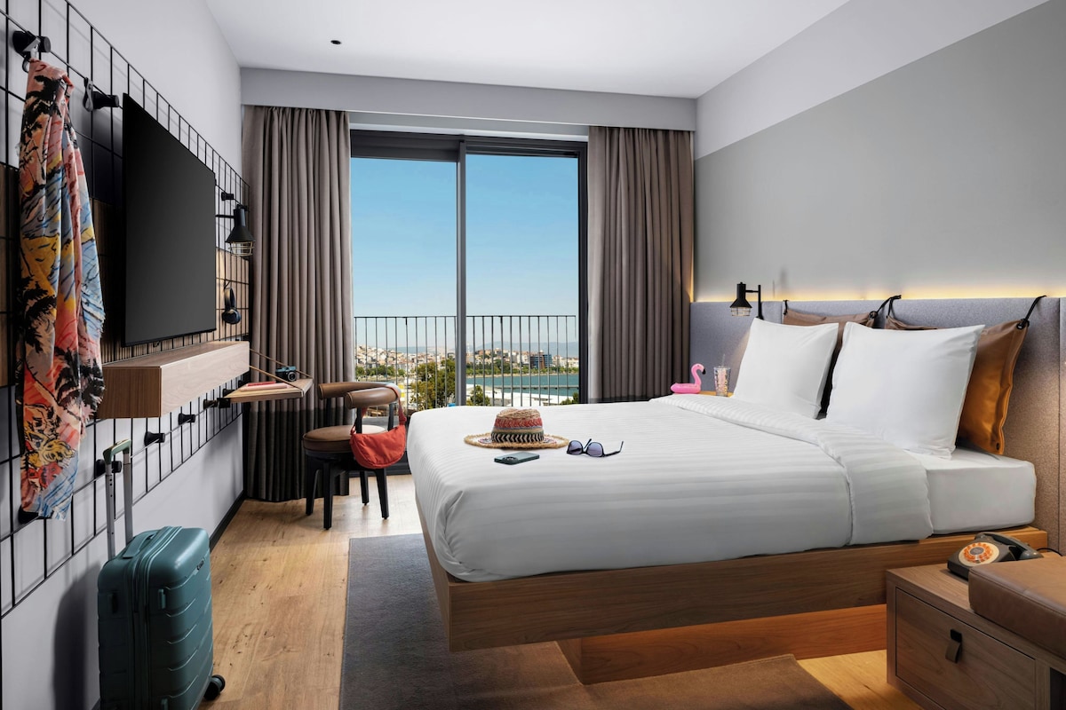A modern hotel room with a large bed, a desk, a chair, a suitcase, and a view of the city through the window.