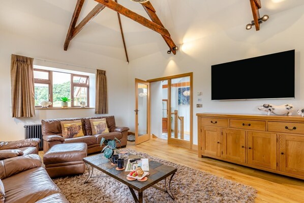 A spacious living room with a brown leather sofa, a coffee table with a plate of food, a wooden cabinet, and a large flat-screen TV. - 3 Bed in Tremar Coombe (oc-g45783) (Tremar)