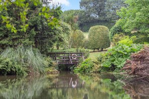 A serene garden with a wooden bridge over a pond, surrounded by lush greenery and various plants. - 3 Bed in Tremar Coombe (oc-g45783) (Tremar)