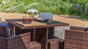 Een tuinmeubelset van riet met een glazen tafel, twee stoelen, een vaas met bloemen en twee glazen rode drank.