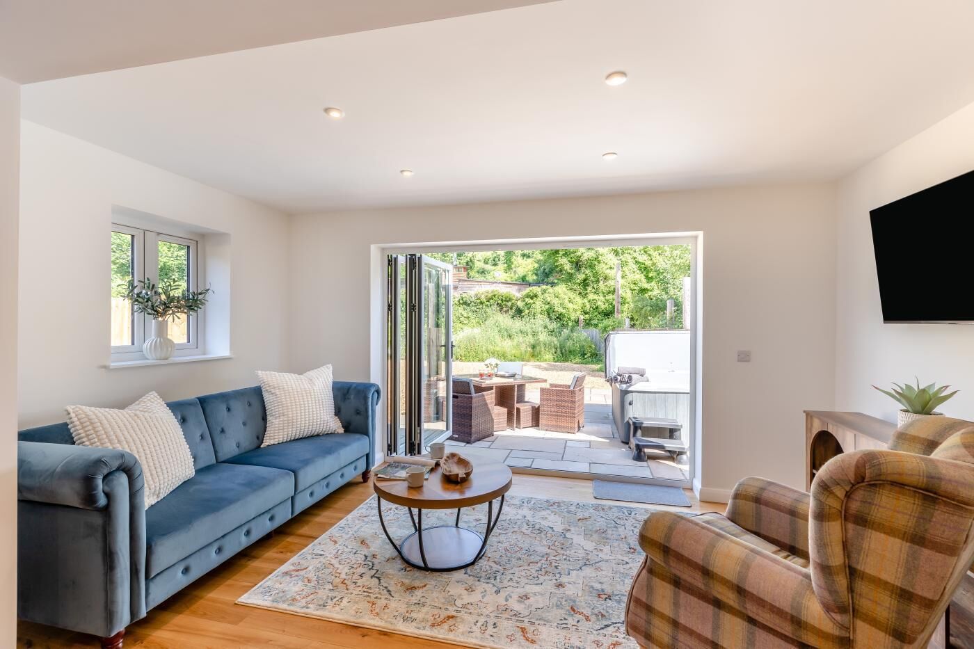 A living room with a blue sofa, a plaid armchair, a coffee table, and a sliding glass door leading to an outdoor patio with a barbecue and garden furniture.
