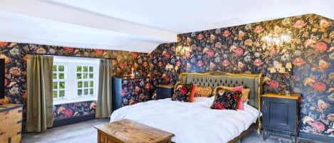 A bedroom with a large bed, floral wallpaper, a wooden nightstand, and a window with curtains.
