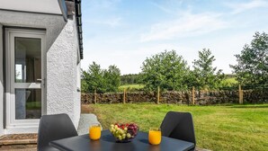 Een tafel met een fruitschaal en twee glazen sinaasappelsap, voor een stenen muur en een grasveld.
