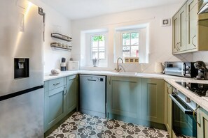 Eine kompakte Küche mit Kühlschrank, Geschirrspüler und Mikrowelle. Zwei Fenster sorgen für Tageslicht, und der Boden ist mit einem gemusterten Fliesenbelag versehen.