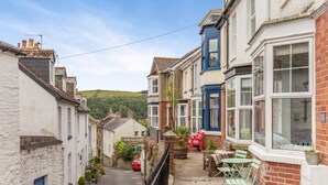 A narrow street with terraced houses, a red car parked on the side, and a small outdoor seating area with a table and chairs. - 3 Bed in Fowey (oc-m32343) (Fowey)