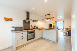 A modern kitchen with a built-in oven, microwave, and a range hood. There is a wooden countertop and a dining area visible in the background. - 2 Bed in Kirkby-in-Furness (oc-s42634) (Kirkby Ireleth)