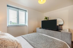 Makuuhuone, jossa on sänky, jalustalla oleva televisio, ikkuna, jossa on sälekaihtimet, ja huonekasvi.