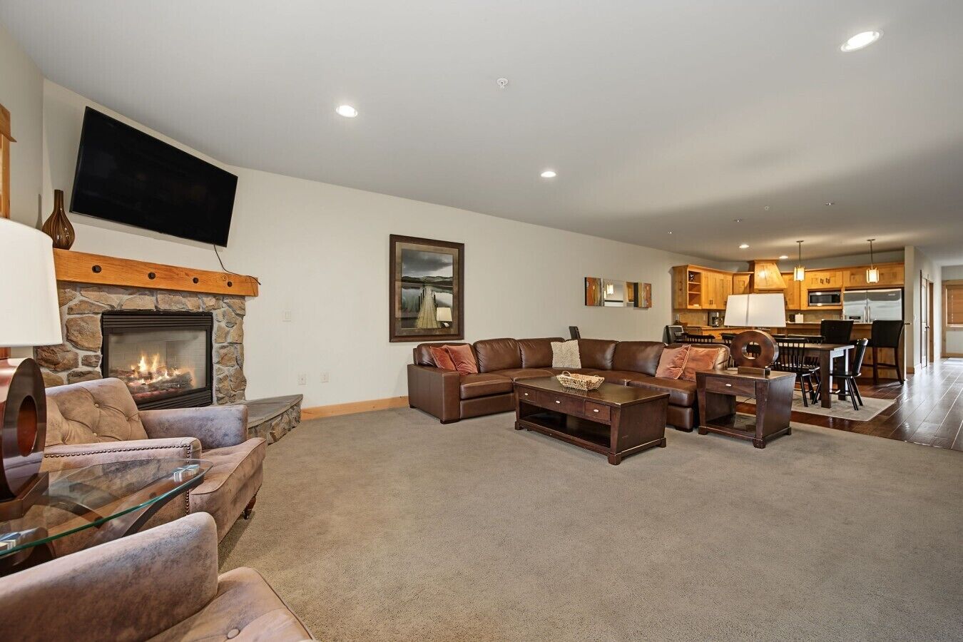 A living room with a stone fireplace, a large flat-screen TV, a sectional sofa, and a dining area with wooden cabinets.