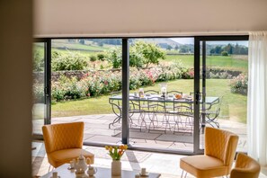 A room with a large sliding glass door leading to an outdoor patio with a table set for dining, two chairs, and a view of a garden and hills. - 4 Bed in Wensley (oc-t45784) (Wensley)