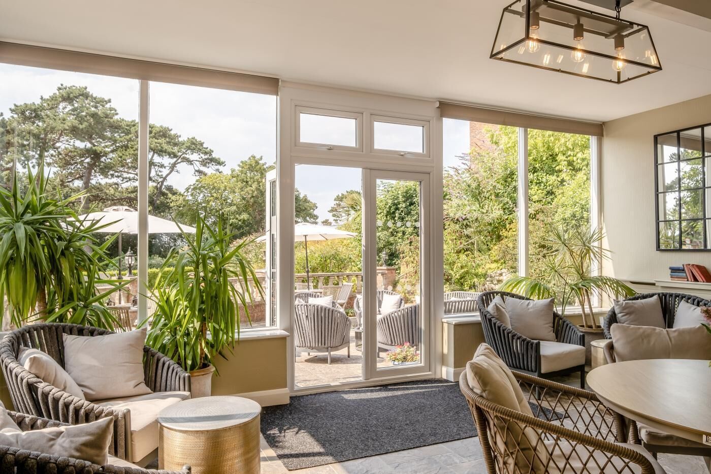 A sunlit room with wicker furniture, a round table, and a view of the outdoors through large windows.