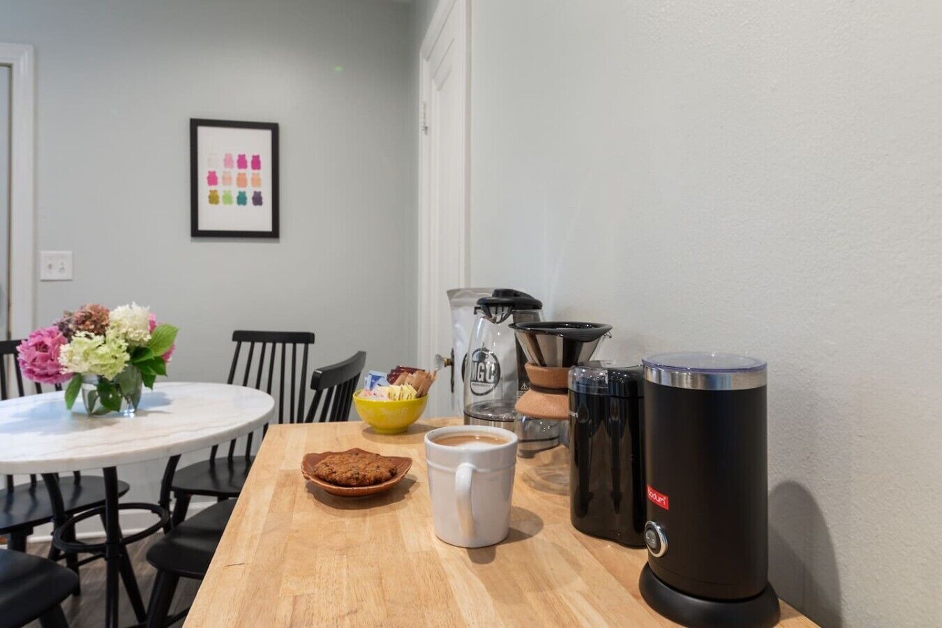 A dining table with a coffee pot, a mug, a bowl of snacks, and a flower vase.