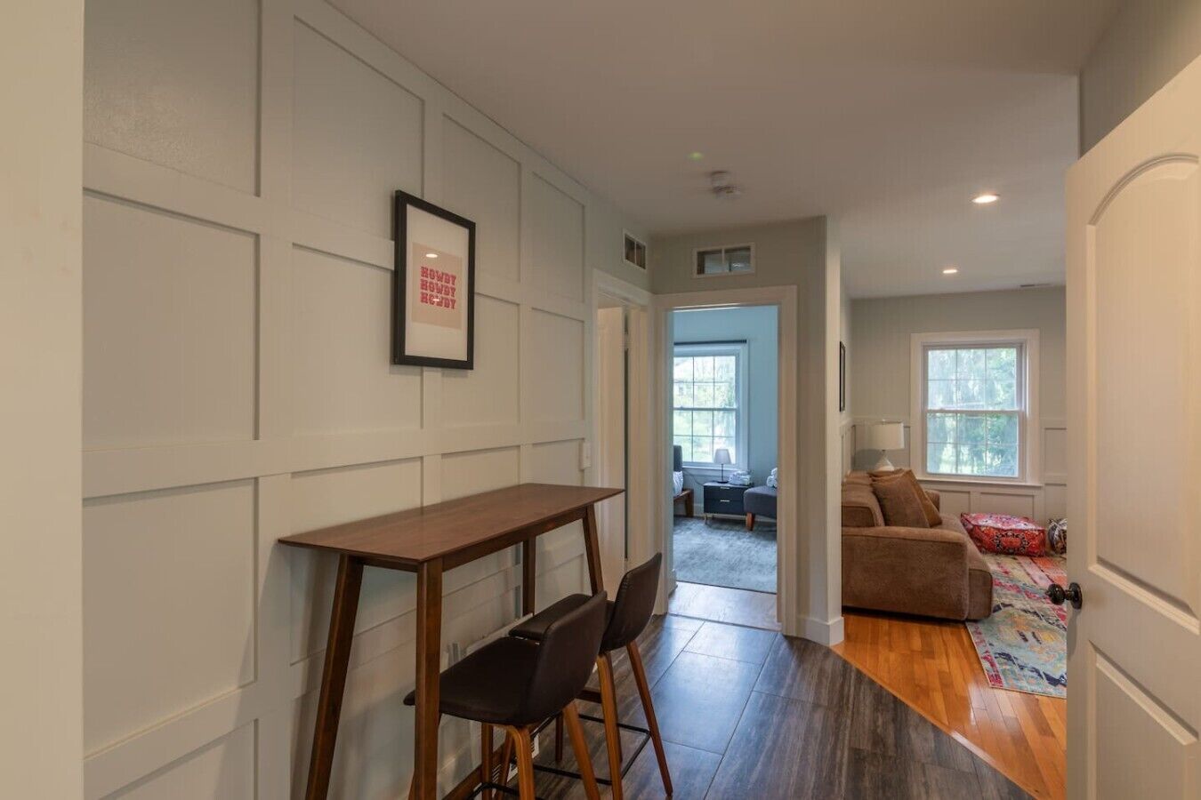 A hallway with a wooden table and chairs, a framed picture on the wall, and a couch in the adjacent room.