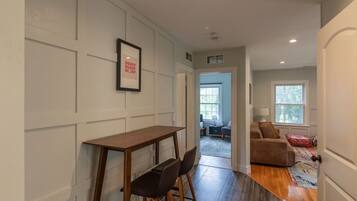 A hallway with a wooden table and chairs, a framed picture on the wall, and a couch in the adjacent room.
