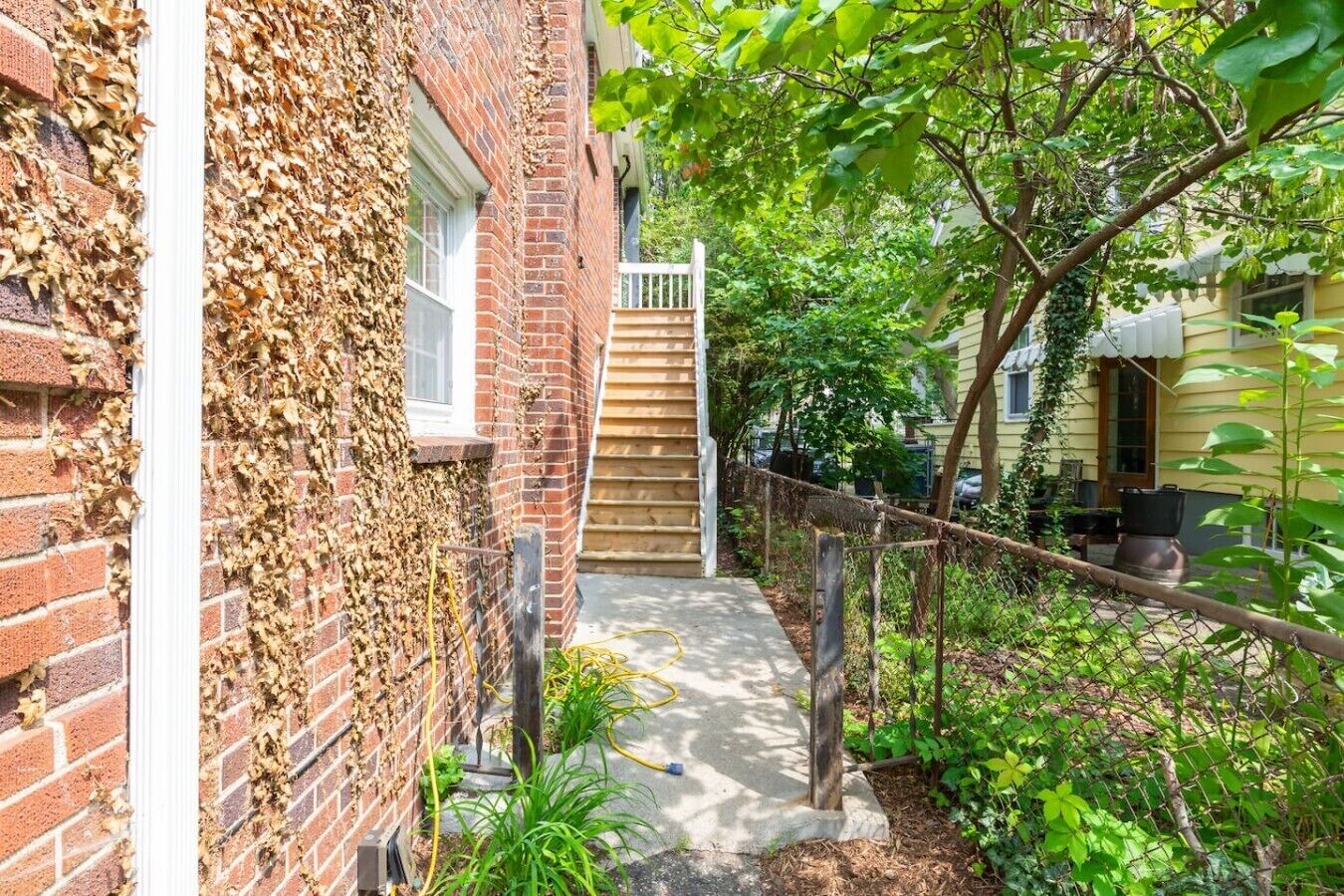 A brick building with a wooden staircase leading to a door, surrounded by greenery and a chain-link fence.