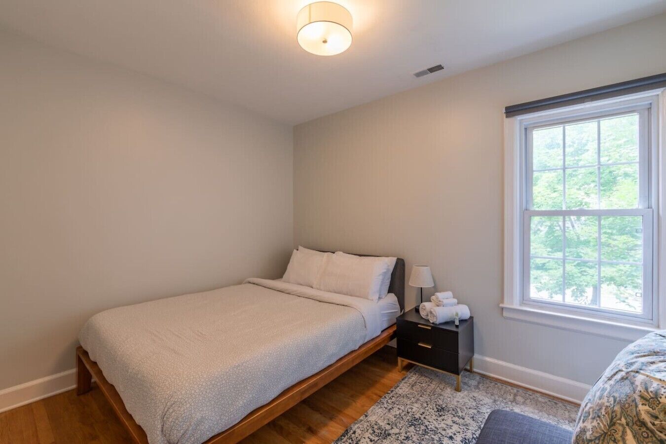 A bedroom with a single bed, a nightstand, a lamp, and a window with a view of trees.