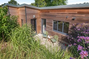 A modern building with a wooden exterior, a patio with chairs, and a garden with tall grass and purple flowers. - 1 Bed in Frampton Mansell (oc-t45435) (Frampton Mansell)