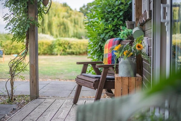 A wooden bench with a colorful blanket and a metal pot with flowers, positioned on a wooden deck.