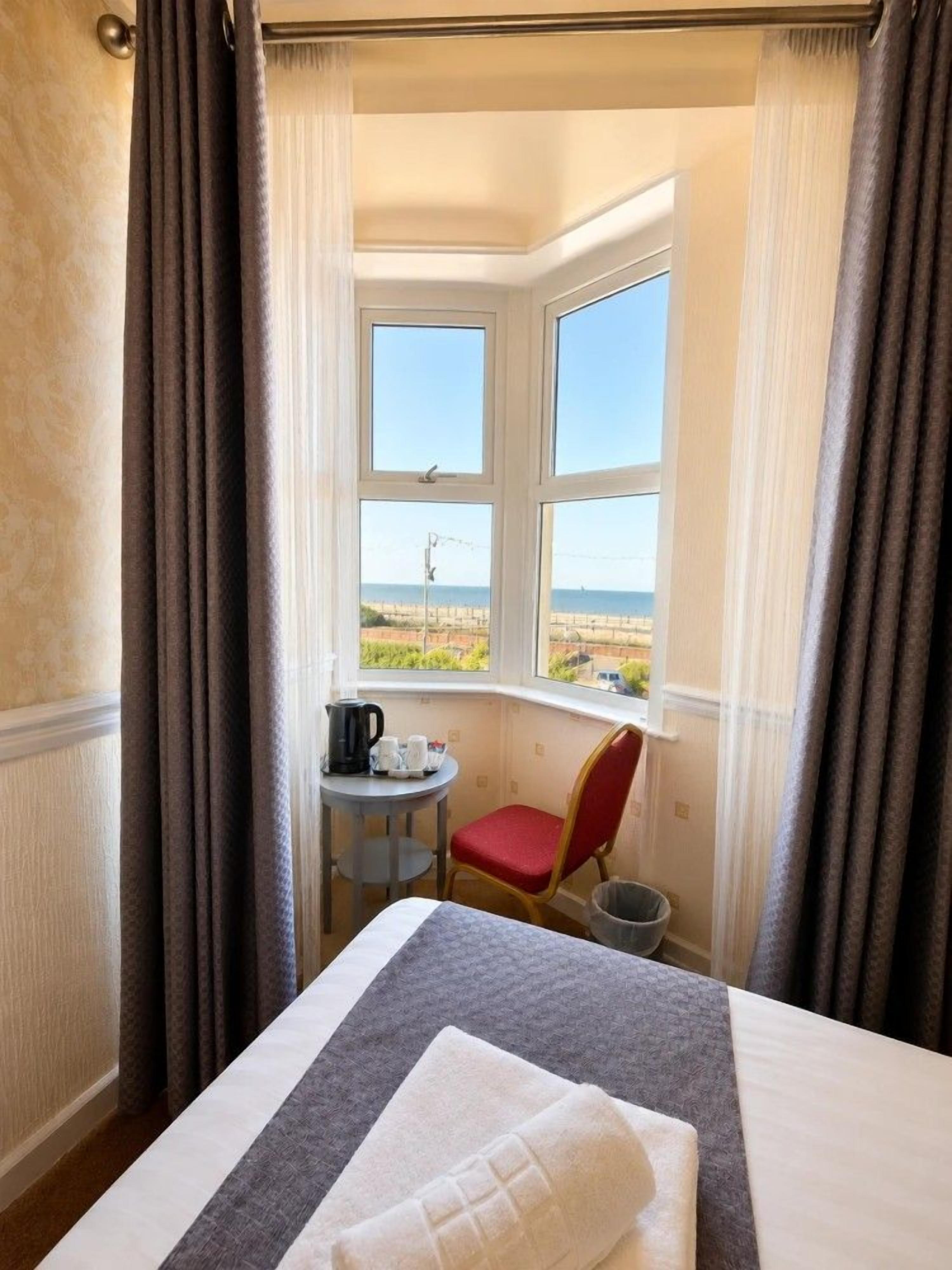 Habitación de hotel con cama, una mesita con tetera y tazas, una silla roja y vista al mar a través de una ventana.