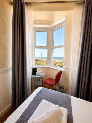 Habitación de hotel con cama, una mesita con tetera y tazas, una silla roja y vista al mar a través de una ventana.