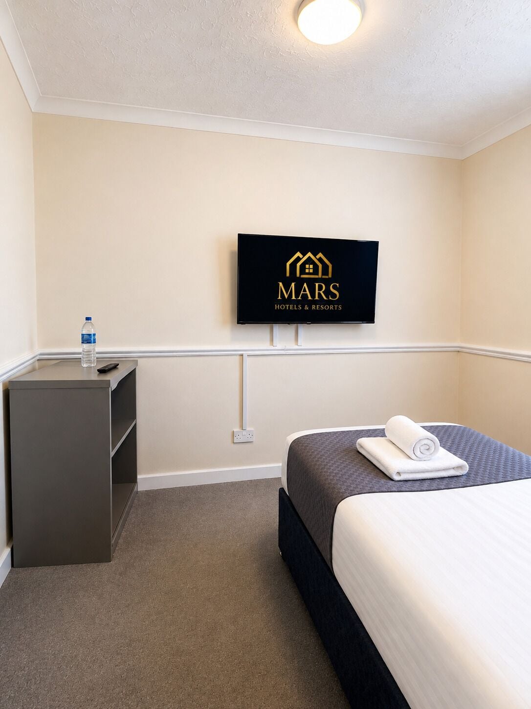 Ett hotellrum med en säng, ett skrivbord, en vattenflaska och en tv som visar hotellets logotyp.