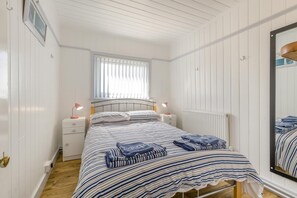 A bedroom with a bed, bedside tables, a window with blinds, a radiator, and a mirror. - 3 Bed in Heacham (oc-v45689) (The Beach)