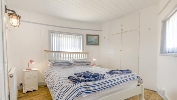 A bedroom with a bed, bedside lamps, a window with blinds, a radiator, and a wardrobe. - 3 Bed in Heacham (oc-v45689) (The Beach)