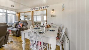 A dining area with a white table set for four, surrounded by white chairs. A large mirror reflects the room, and a 'VIVE' banner hangs above the table. - 3 Bed in Heacham (oc-v45689) (The Beach)